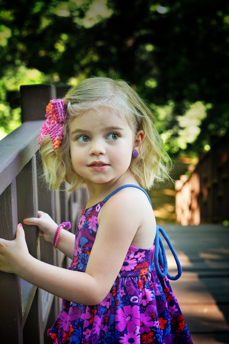 May include: A young girl with blonde hair wearing a purple and pink floral dress with a blue strap. She is wearing a pink flower in her hair and purple earrings. She is standing next to a wooden railing.