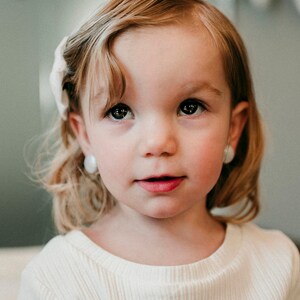 May include: A young girl with blonde hair and a white shirt looks directly at the camera. She is wearing small white earrings.