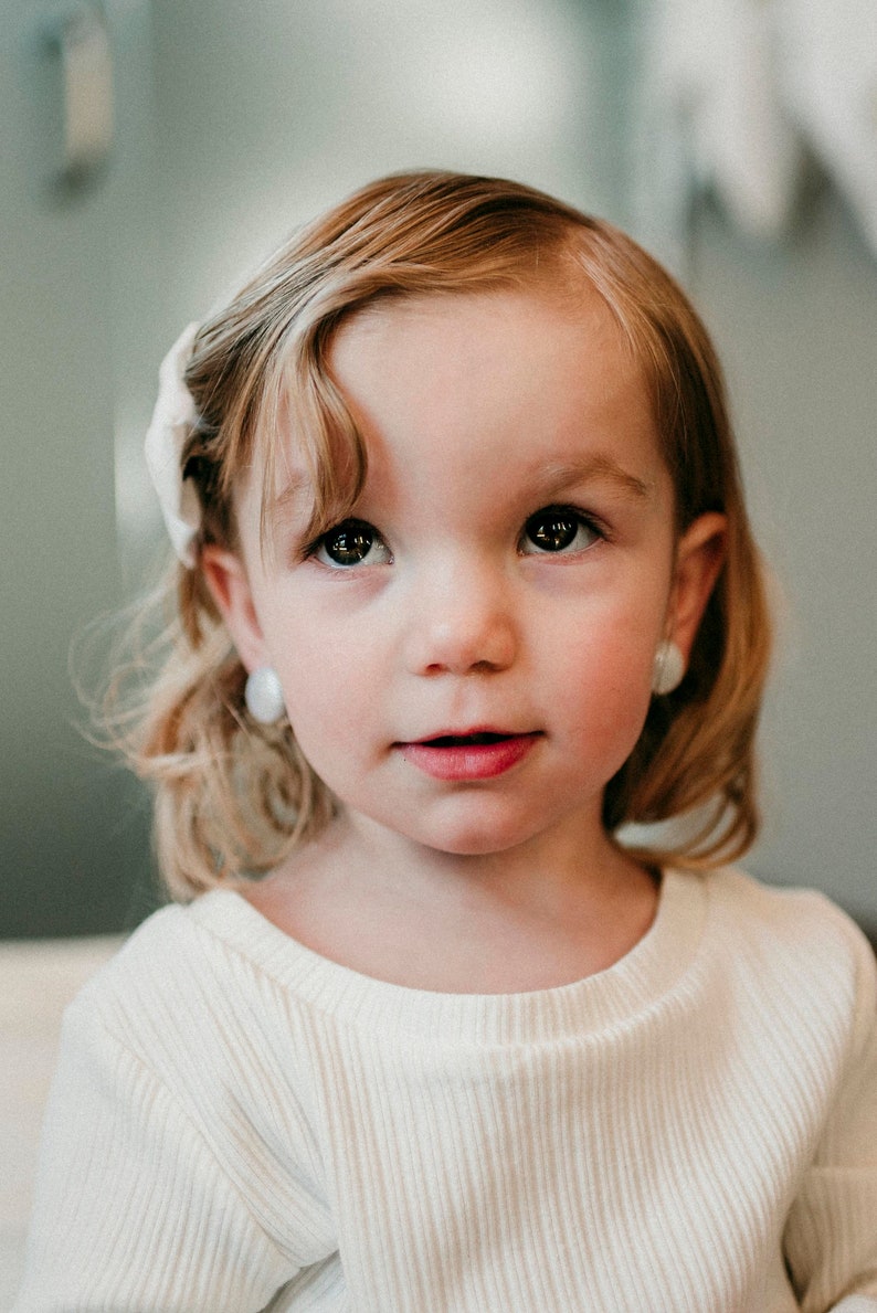 May include: A young girl with blonde hair and a white shirt looks directly at the camera. She is wearing small white earrings.