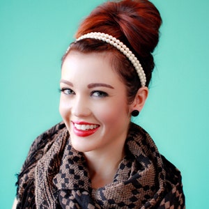 May include: A woman with red hair styled in a high bun wears a pearl headband and a black and brown patterned scarf. She is smiling with red lipstick.