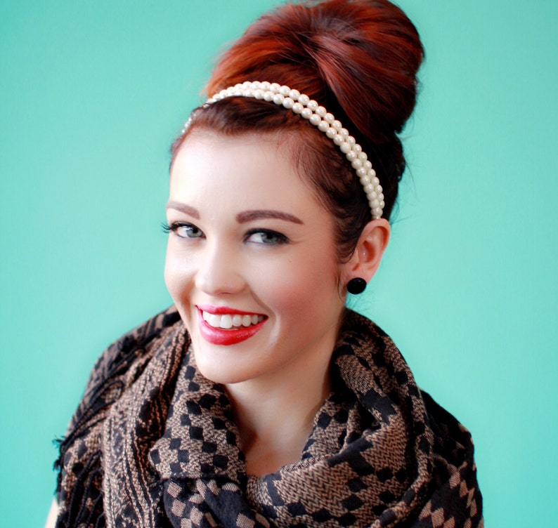 May include: A woman with red hair styled in a high bun wears a pearl headband and a black and brown patterned scarf. She is smiling with red lipstick.