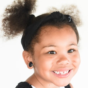 May include: A young girl with curly black hair is wearing a black headband and a black and white striped shirt. She is smiling and has a gap in her front teeth.