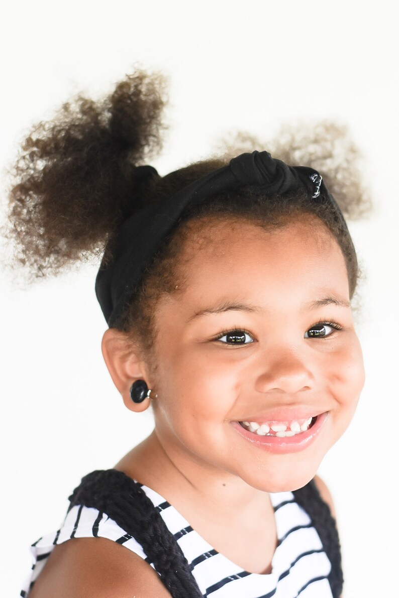 May include: A young girl with curly black hair is wearing a black headband and a black and white striped shirt. She is smiling and has a gap in her front teeth.