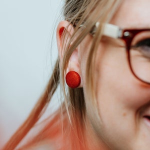 May include: A close-up of a person's ear with a red button-shaped earring. The person is wearing glasses.