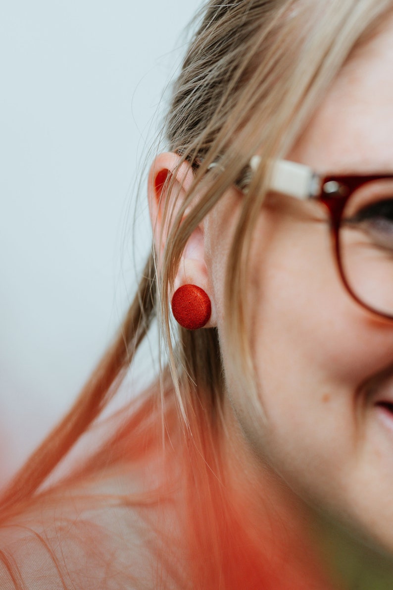 May include: A close-up of a person's ear with a red button-shaped earring. The person is wearing glasses.