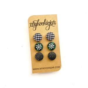 May include: Three pairs of circular fabric-covered stud earrings on a tan display card labelled mykookiejar. The top pair features a black and white buffalo check pattern, the middle pair has a green plaid print with white snowflakes, and the bottom pair is solid black with gold glitter flecks.