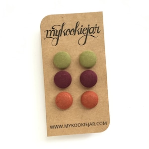 Puede incluir: Conjunto de seis pendientes de botón de tela en tres colores: verde, burdeos y naranja. Los pendientes se muestran en una tarjeta marrón con el texto "mycookiejar" y "www.mycookiejar.com".