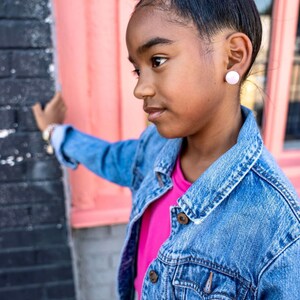 May include: A young person with dark brown hair in a bun, wearing a pink t-shirt and a blue denim jacket, is standing in front of a pink wall. They are looking to the left of the frame and have a white button earring in their left ear.