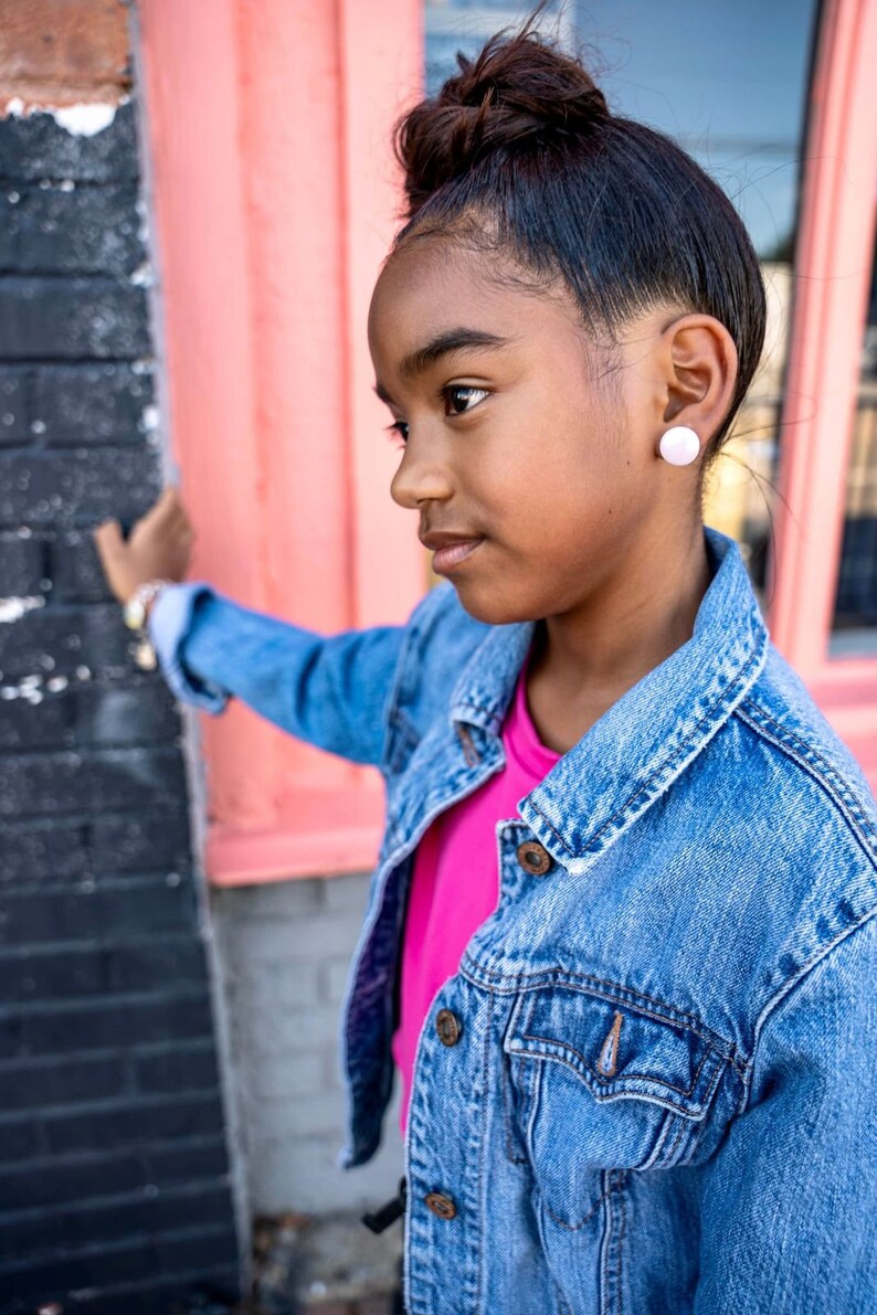 May include: A young person with dark brown hair in a bun, wearing a pink t-shirt and a blue denim jacket, is standing in front of a pink wall. They are looking to the left of the frame and have a white button earring in their left ear.