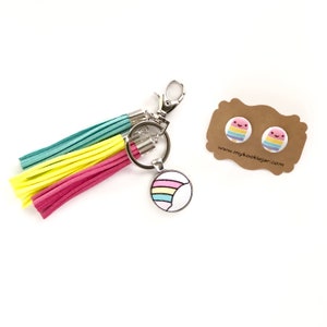 May include: A rainbow-colored keychain with a tassel and a round charm featuring a rainbow design. The keychain is accompanied by a pair of rainbow-colored earrings with a smiling face design. The earrings are on a brown card with the text "www.mykookiejjar.com".