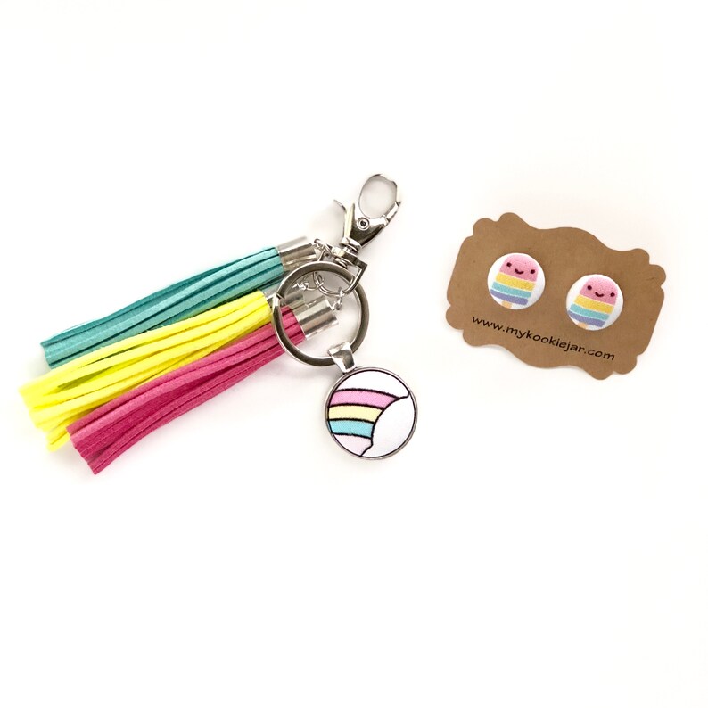 May include: A rainbow-colored keychain with a tassel and a round charm featuring a rainbow design. The keychain is accompanied by a pair of rainbow-colored earrings with a smiling face design. The earrings are on a brown card with the text "www.mykookiejjar.com".