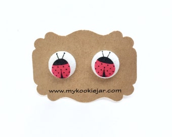 Könnte beinhalten: Ein Paar Ohrringe aus Stoff in Form eines Marienkäfers auf einer braunen Karte mit der Website www.mycookiejar.com.