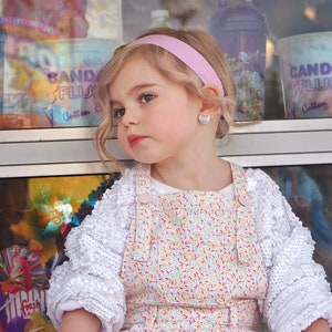 May include: A young girl with blonde hair wearing a white shirt with sequins and a pink and white overall dress with a sprinkle pattern. She is wearing a pink headband and rainbow-colored earrings. The girl is sitting in front of a window with a sign that says "Candy Fluff Cotton Candy".