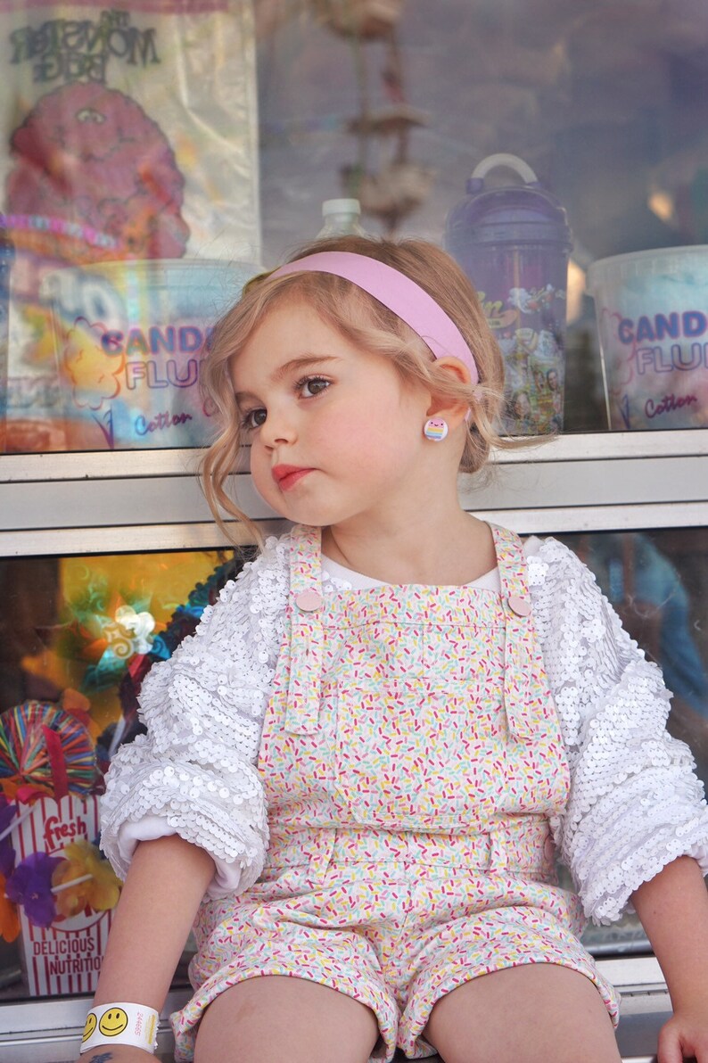 May include: A young girl with blonde hair wearing a white shirt with sequins and a pink and white overall dress with a sprinkle pattern. She is wearing a pink headband and rainbow-colored earrings. The girl is sitting in front of a window with a sign that says "Candy Fluff Cotton Candy".