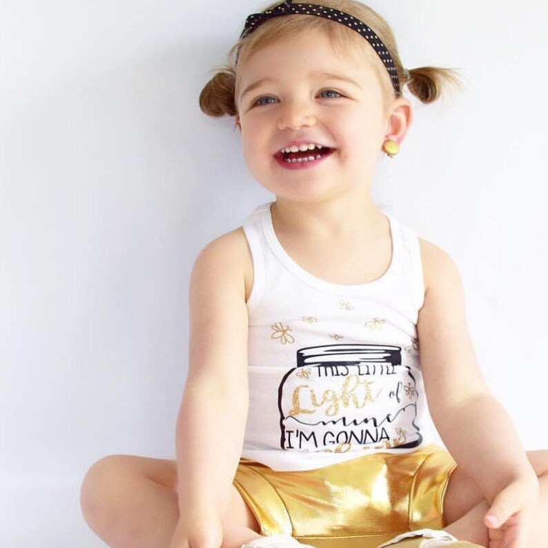May include: A white tank top with a gold glitter design of a mason jar with the text "This little light of mine I'm gonna let it shine". The tank top is being worn by a young child with pigtails and a black polka dot headband. The child is wearing gold shorts.