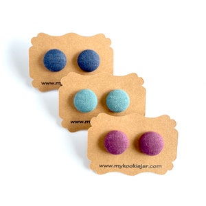 May include: Three pairs of fabric button earrings on brown card stock. The earrings are in shades of blue, teal, and purple.