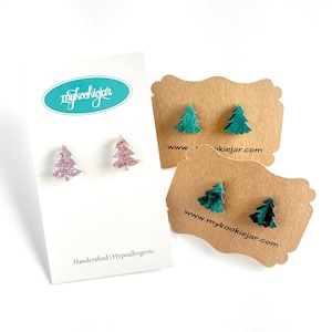 Puede incluir: Pendientes con forma de árboles de Navidad. Un par es de color rosa claro con purpurina, y los otros dos pares son de color verde azulado y verde azulado oscuro. Los pendientes se muestran en tarjetas blancas y marrones. La tarjeta blanca tiene el texto "mykookiejar".