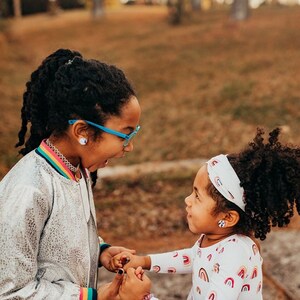 May include: Two young girls with curly dark hair are smiling and holding hands. The girl on the left is wearing a silver jacket with a rainbow stripe and blue glasses. The girl on the right is wearing a white shirt with a rainbow pattern and a white headband with a rainbow pattern.