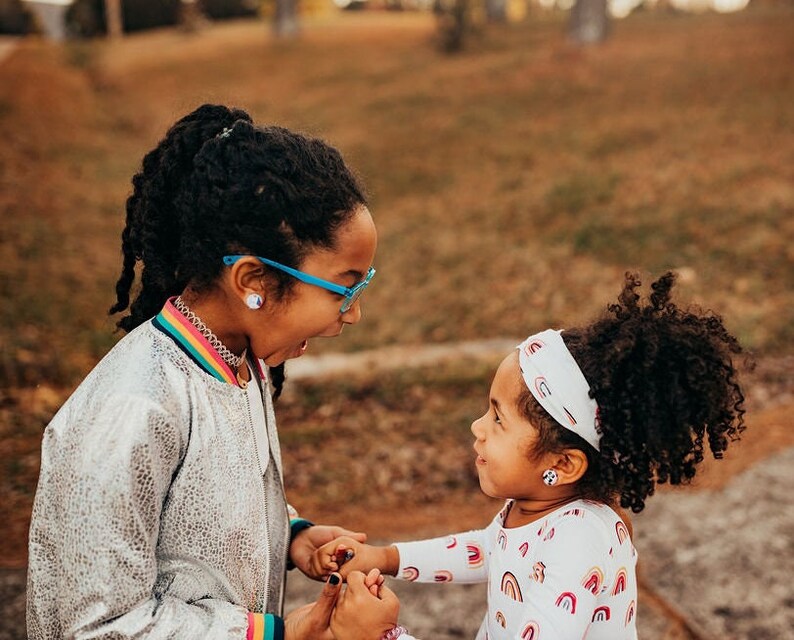 May include: Two young girls with curly dark hair are smiling and holding hands. The girl on the left is wearing a silver jacket with a rainbow stripe and blue glasses. The girl on the right is wearing a white shirt with a rainbow pattern and a white headband with a rainbow pattern.
