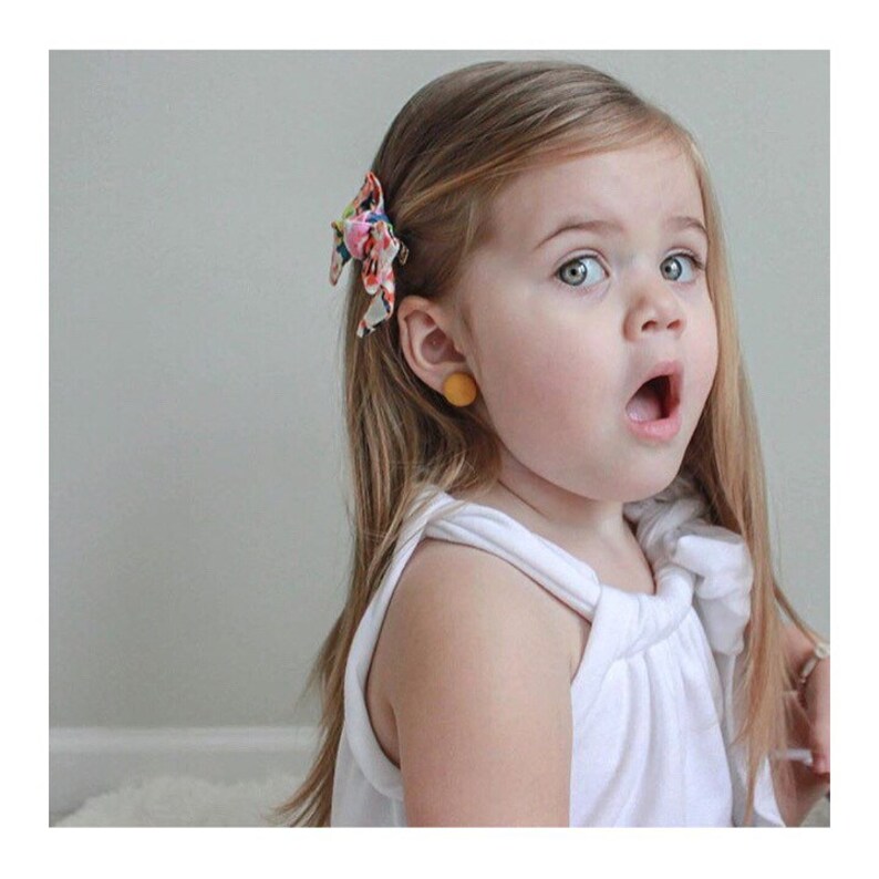 May include: A young girl with long blonde hair wearing a white tank top and a floral hair clip. She is wearing a yellow round earring in her left ear.