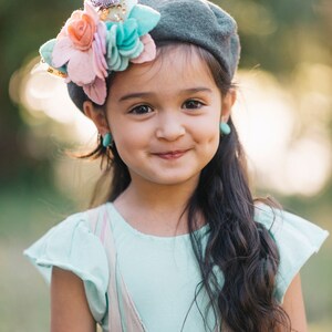 May include: A young girl with long dark hair smiles while wearing a gray beret with a pink, blue, and white flower embellishment. She is wearing a light blue shirt with a pink and white striped overall dress.