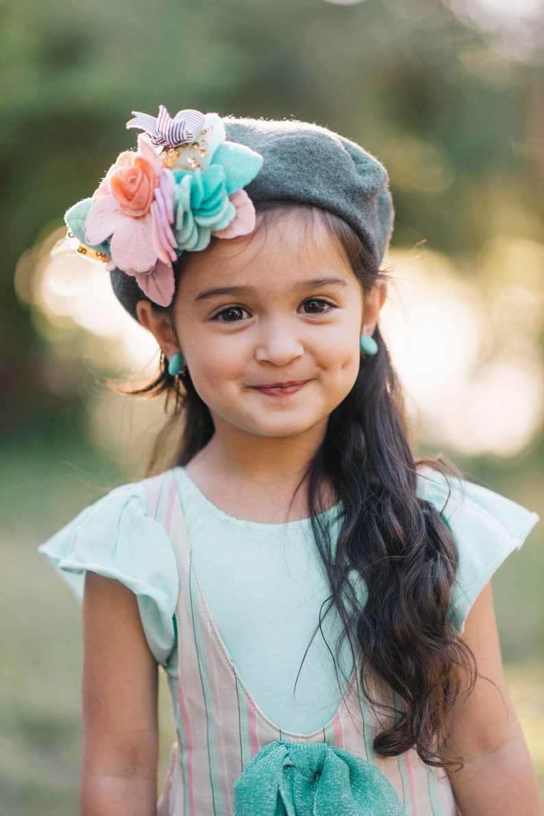 May include: A young girl with long dark hair smiles while wearing a gray beret with a pink, blue, and white flower embellishment. She is wearing a light blue shirt with a pink and white striped overall dress.