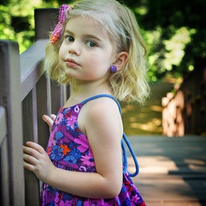 May include: A young girl with blonde hair wearing a purple and blue floral sundress with a blue strap. She is standing next to a wooden fence and has a pink bow in her hair.
