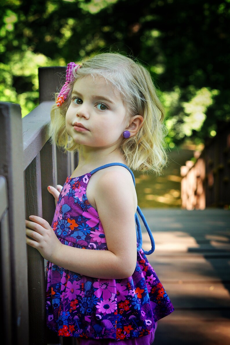 May include: A young girl with blonde hair wearing a purple and blue floral sundress with a blue strap. She is standing next to a wooden fence and has a pink bow in her hair.