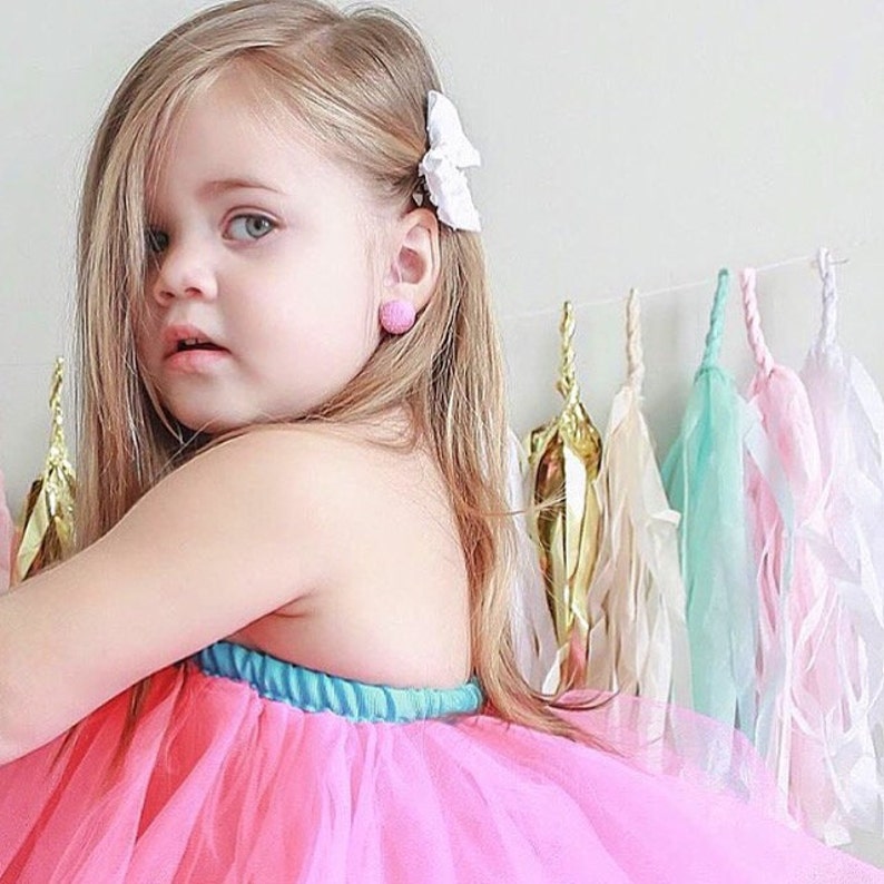 May include: A young girl with long blonde hair wearing a pink tulle tutu skirt and a white bow in her hair. She is wearing pink earrings and has a turquoise and blue trim on her skirt.