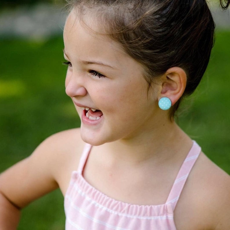 May include: A young girl with brown hair wearing a pink and white striped tank top and light blue polka dot earrings. She is smiling and looking to the side.