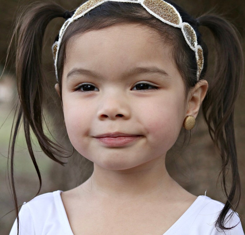 May include: A young girl with long brown hair wearing a gold and white headband with a sparkly design. She is wearing a white tank top and has a gold stud earring in her left ear.