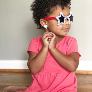 May include: A young girl with curly hair wearing a pink dress and star-shaped sunglasses with black polka dots.