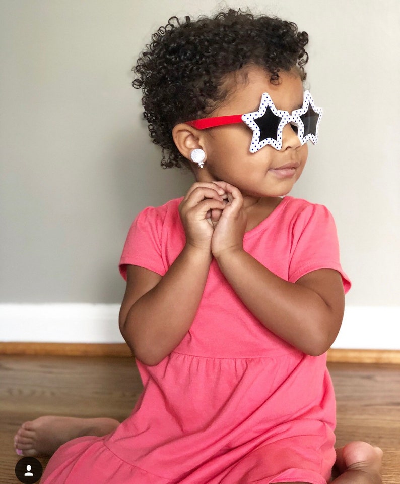 May include: A young girl with curly hair wearing a pink dress and star-shaped sunglasses with black polka dots.