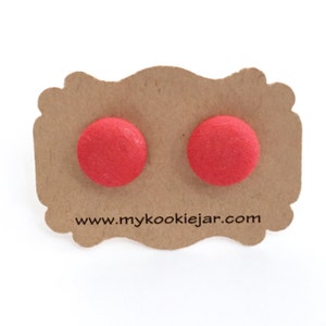 May include: A pair of round, bright red fabric-covered stud earrings attached to a scalloped brown cardboard display card. The card features the printed text www.mykookiejar.com centered at the bottom below the two circular red earrings.