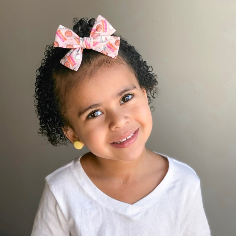May include: A young girl with curly brown hair wearing a white t-shirt and a pink bow with a donut pattern. She is wearing yellow earrings.