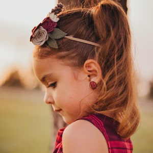 May include: A young girl with long brown hair wearing a red and black plaid dress and a flower headband. She is wearing red glitter earrings.