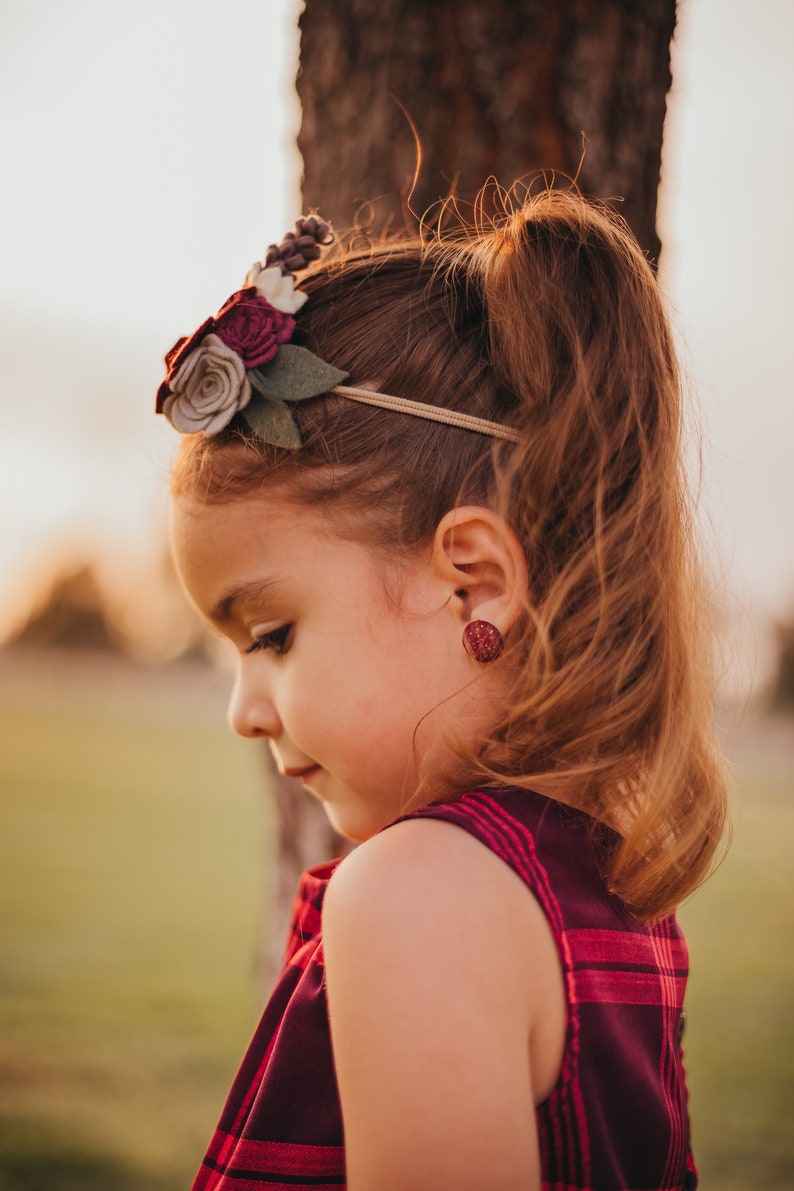 May include: A young girl with long brown hair wearing a red and black plaid dress and a flower headband. She is wearing red glitter earrings.