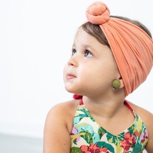 May include: A young girl with brown hair wearing a coral turban headband and a green wooden earring. She is wearing a floral print halter top.