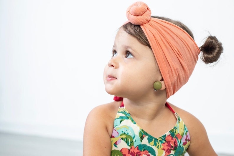 May include: A young girl with brown hair wearing a coral turban headband and a green wooden earring. She is wearing a floral print halter top.