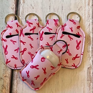 May include: Pink hand sanitizer holders with a repeating pattern of pink ribbons, each attached to a silver keyring. The holders are designed to carry a small hand sanitizer bottle. The holders are a light pink color.