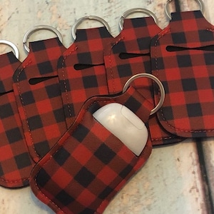 May include: A collection of red and black plaid fabric keychains. One keychain holds a white earbud case. The keychains have a silver ring and a small opening for access.
