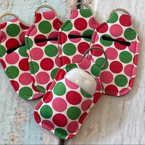 May include: A collection of five polka-dot hand sanitizer holders with keychains. The holders feature a pattern of red, green, and pink circles on a white background. Each holder has a black opening for the sanitizer bottle and a gold keychain ring.