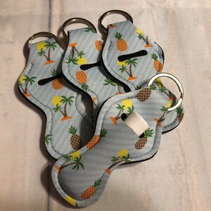 May include: Four blue and white lip balm holders with pineapple and palm tree designs. Each holder has a silver key ring.