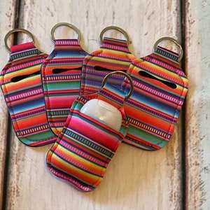 May include: Six colorful striped hand sanitizer holders with keychains. The holders are made of a soft, flexible material and have a small opening for a hand sanitizer bottle. The holders are all in a similar pattern, with stripes of red, orange, yellow, green, blue, and purple.