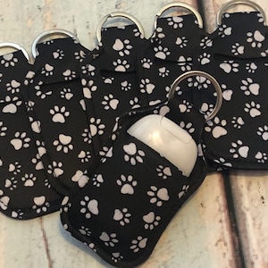 May include: Six black fabric keychains with white paw print designs. Each keychain has a silver ring and a clear plastic bottle holder.