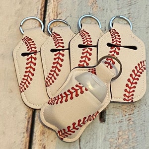 May include: Baseball-themed hand sanitizer holders with keychains. The holders are off-white with red stitching, resembling baseballs. The design includes a slot for the sanitizer bottle and a metal keyring.