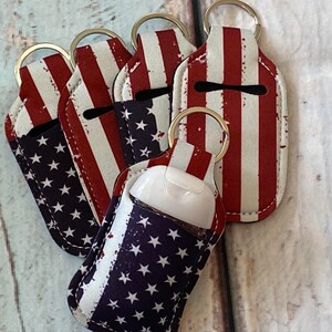 May include: Several hand sanitizer holders with key rings. The holders are decorated with a distressed American flag pattern of red and white stripes and a blue field of white stars. A small, white hand sanitizer bottle is visible in one holder.