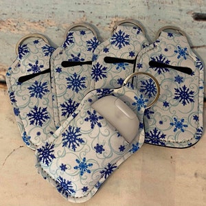 May include: Six white hand sanitizer holders with blue snowflake patterns and silver keychains. The holders are made of soft, flexible material and are designed to fit a standard-sized hand sanitizer bottle.