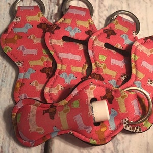 May include: Five pink keychains with a dachshund pattern and silver key rings. Each keychain has a small pocket for a lip balm or other small item. One keychain has a lip balm inside the pocket.