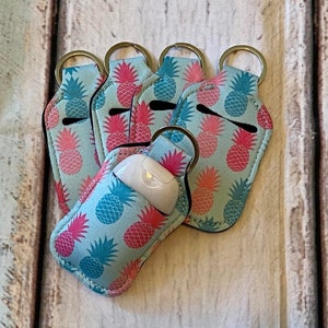 May include: Six teal blue hand sanitizer holders with a pineapple print. The holders have silver key rings and are made of a soft, flexible material.